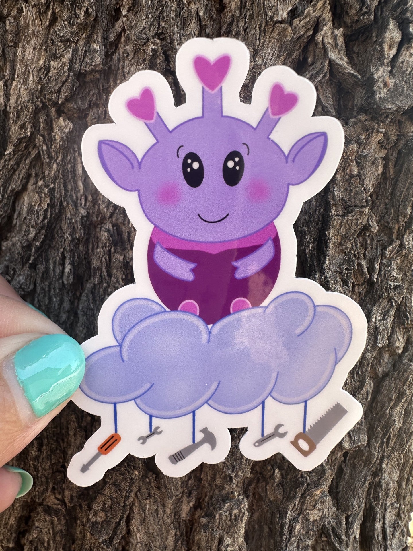 Purple alien cartoon character with a cloud and tools hanging from it. The purple alien has hearts on the top of its head, big black eyes, and rosy cheeks. The sticker is held against a tree trunk.