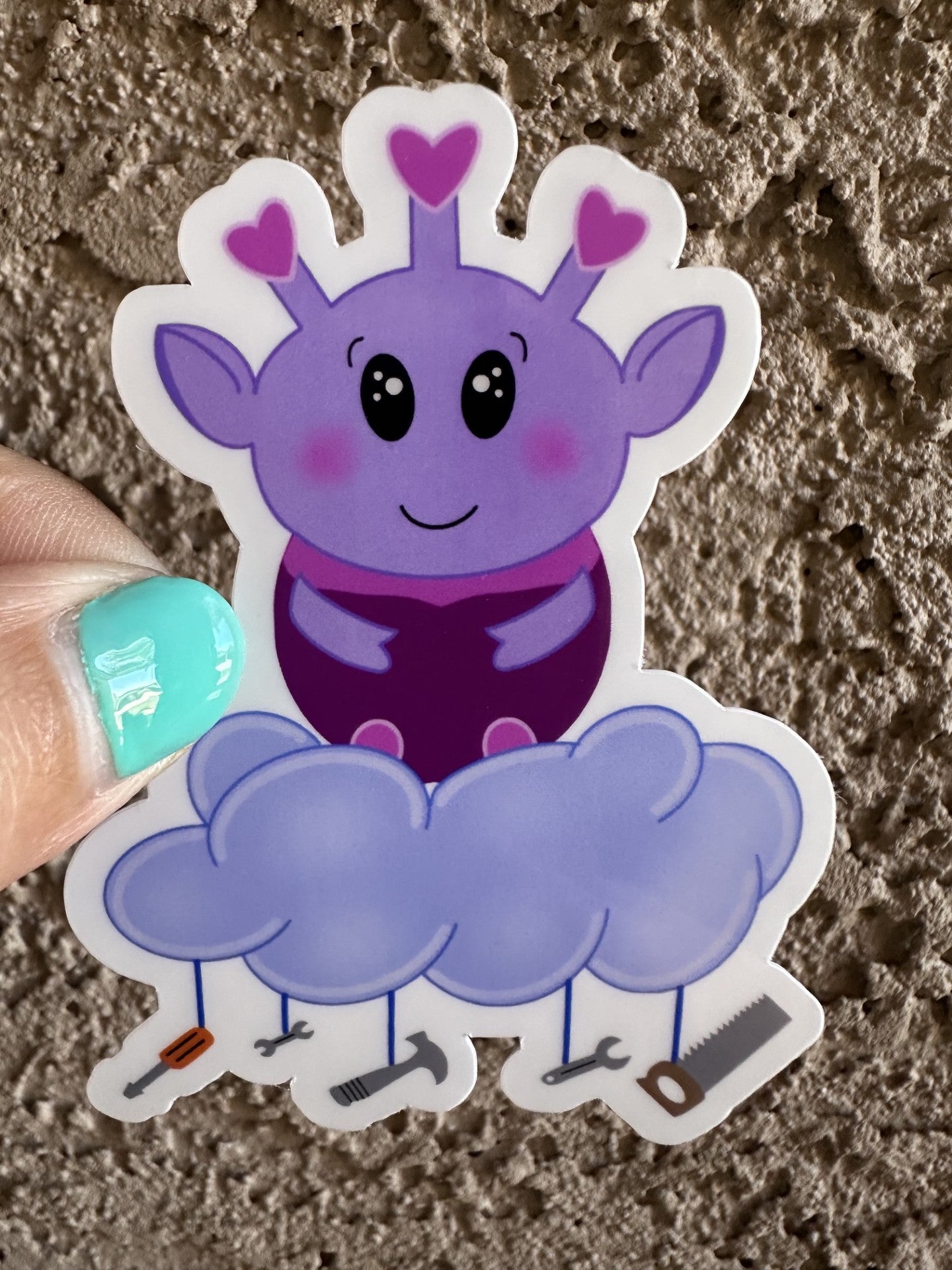 Purple alien cartoon character with a cloud and tools hanging from it. The purple alien has hearts on the top of its head, big black eyes, and rosy cheeks. The sticker is held against a textured surface.