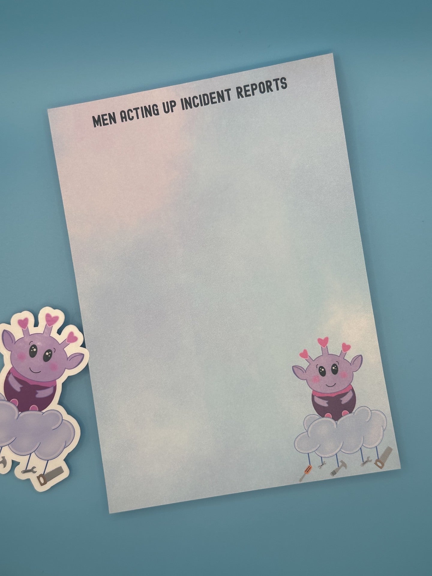 Notepad with a cartoon character at the bottom right of the notepad. The character looks like an alien who is purple and is sitting on top of a cloud with tools hanging from it. The notepad has a pastel-colored background, and on top of the notepad states 'Men Acting Up Incident Reports'. The notepad is sitting on a blue background with a sticker.