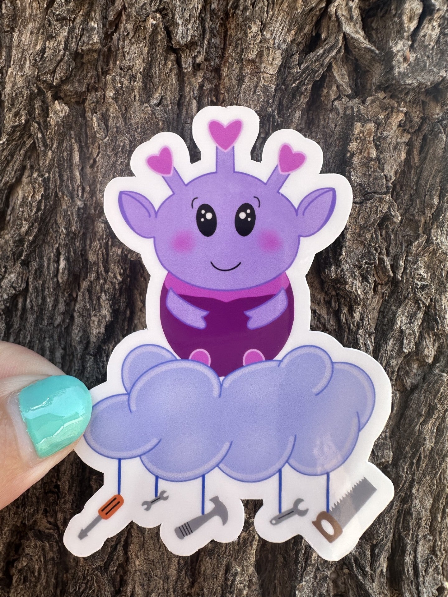 Purple alien cartoon character with a cloud and tools hanging from it. The purple alien has hearts on the top of its head, big black eyes, and rosy cheeks. The sticker is held against a tree trunk.