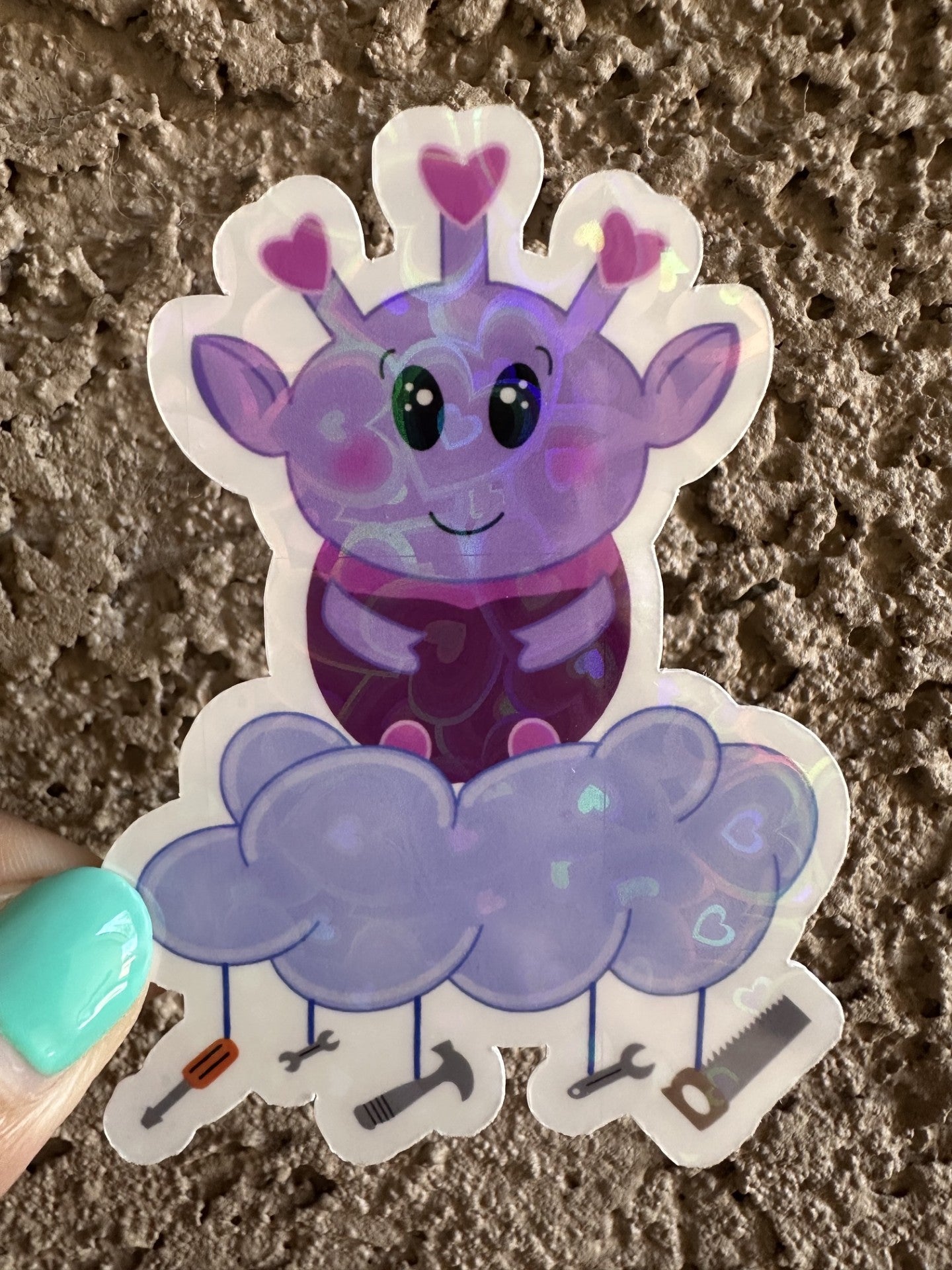 Purple alien cartoon character with a cloud and tools hanging from it. The purple alien has hearts on the top of its head, big black eyes, and rosy cheeks. The heart holographic sticker is held against a textured surface.