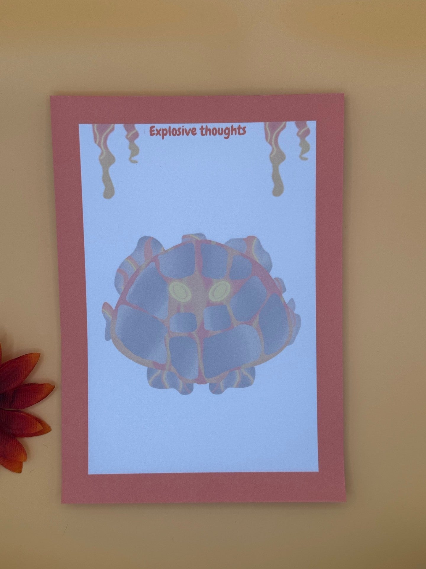 A notepad that has rocks and orange and red lava coming from it. The lava character has yellow eyes. The notepad has orange edges all around the four corners and has lava coming down from the top of the notepad. The notepad has a heading of " Explosive thoughts" The notepad is sitting on top of an orange background with a flower.