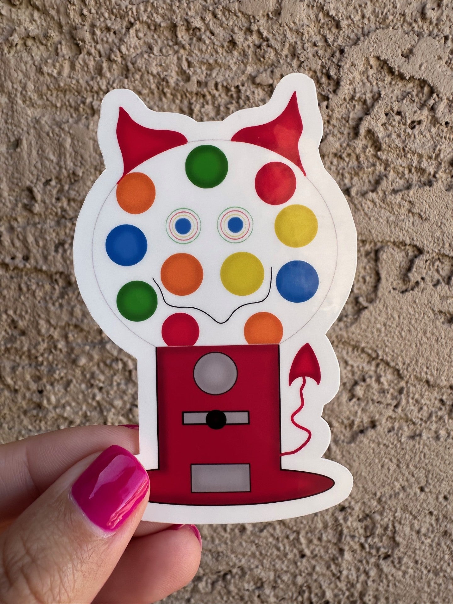 A red gumball machine shaped with different colors of gumballs inside and a black smirk with colorful eyes. The character also has horns and a tail. The sticker is held by a hand against a textured wall.