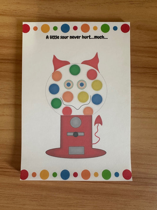Colorful gumball machine illustration with a sneaky smile that has devil horns and a devil tail. The notepad is on a wooden surface.