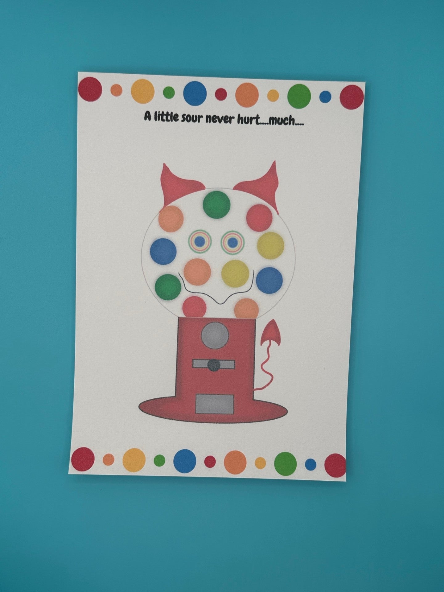 A notepad that has a red gumball machine character. Inside of the gumball machine is different color gumballs and coming out of the character is red horns and a red devil tail. The notepad has red, orange, green and blue dots on the top of the notepad and bottom. The heading of the notepad states "A little sour never hurt....much...". The notepad is sitting on top of a blue background.