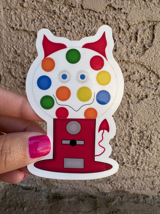 Colorful gumball machine-shaped sticker with horns and a tail held by a hand against a textured wall.