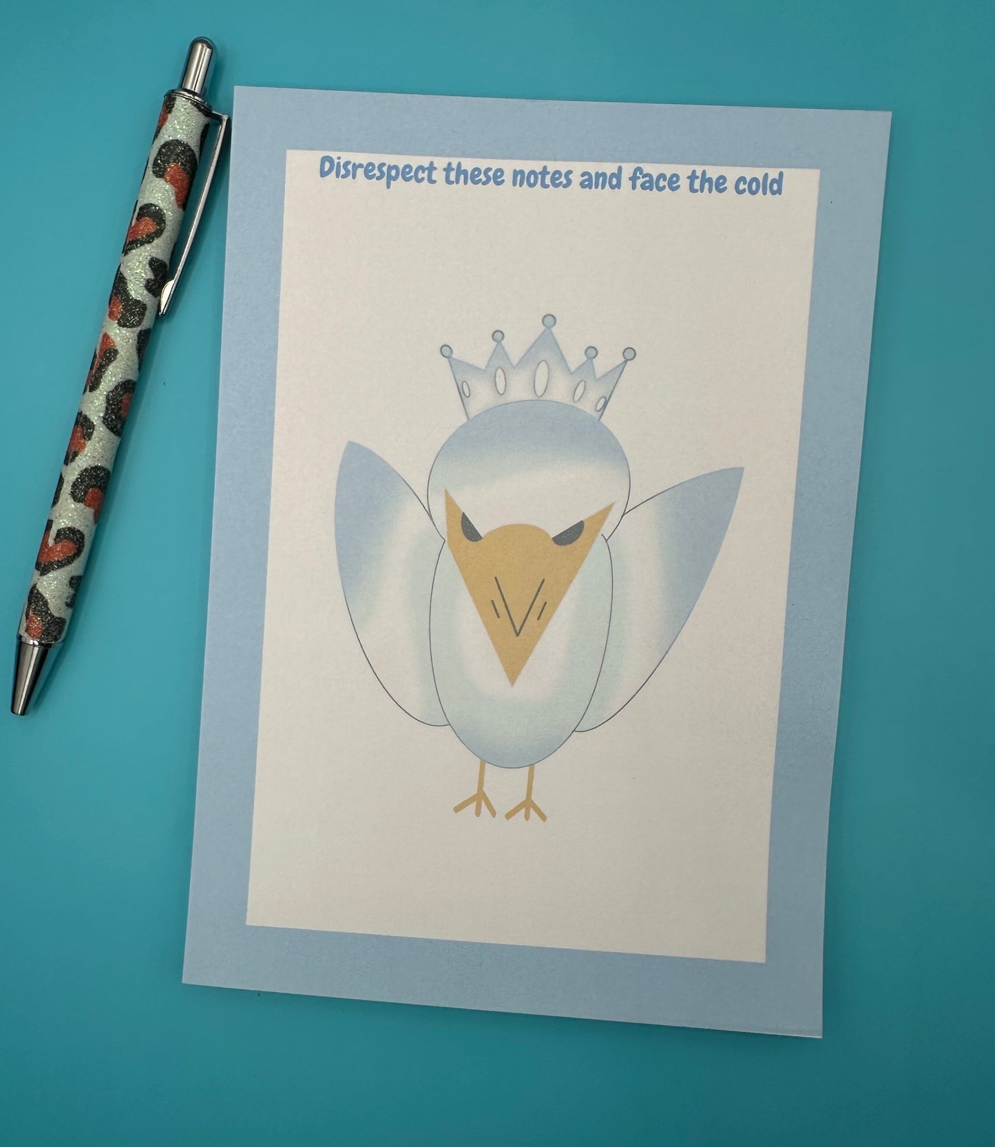 A notepad that has a bird character on it that is blue and white color with a yellow snout and black eyes and also has a crown on top of its head. The notepad edges has a light blue color with the heading on top of the notepad stating "Disrespect these notes and face the cold". The notepad is sitting on top of a blue background with a pen.