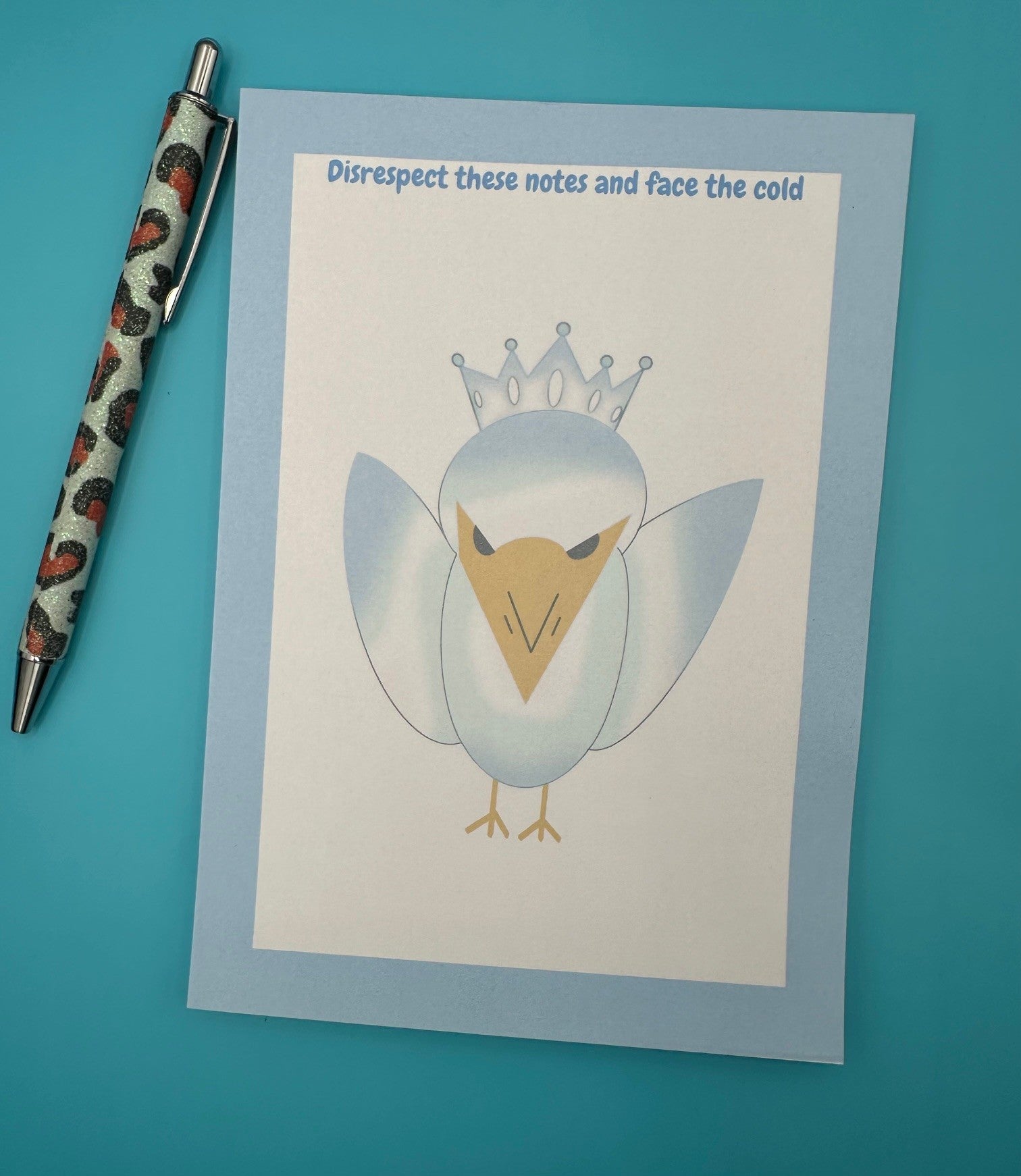 A notepad that has a bird character on it that is blue and white color with a yellow snout and black eyes and also has a crown on top of its head. The notepad edges has a light blue color with the heading on top of the notepad stating "Disrespect these notes and face the cold". The notepad is sitting on top of a blue background with a pen.
