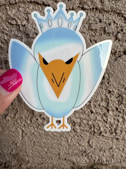 Sticker of a bird character with a crown held by a hand on a textured surface. Colors of the bird is blue and light blue.