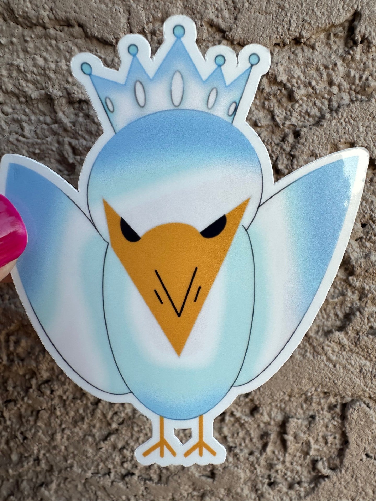 A cartoon bird sticker with blue and light blue colors, with a crown and a yellow snout. The background is on a textured surface.