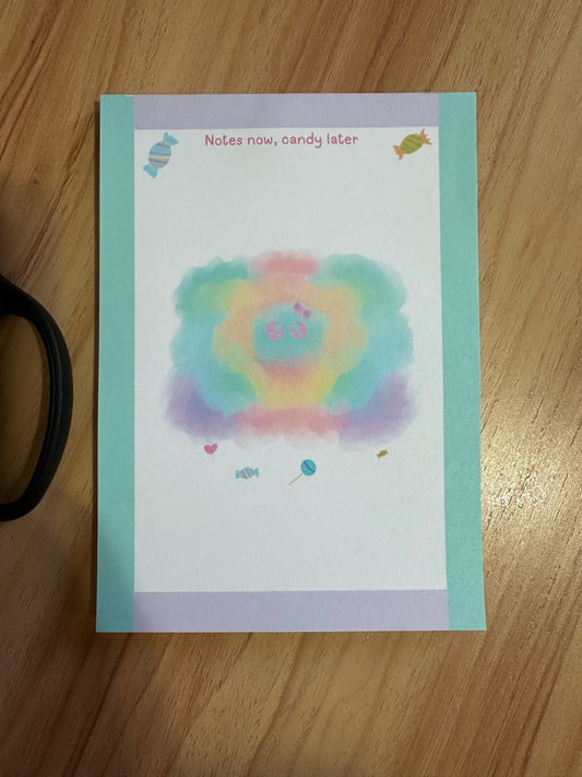 Colorful character design "sprinkles" that has different pastel colors with candy beneath the character. The notepad is sitting on wooden surface with a heading on the notepad "notes now, candy later".
