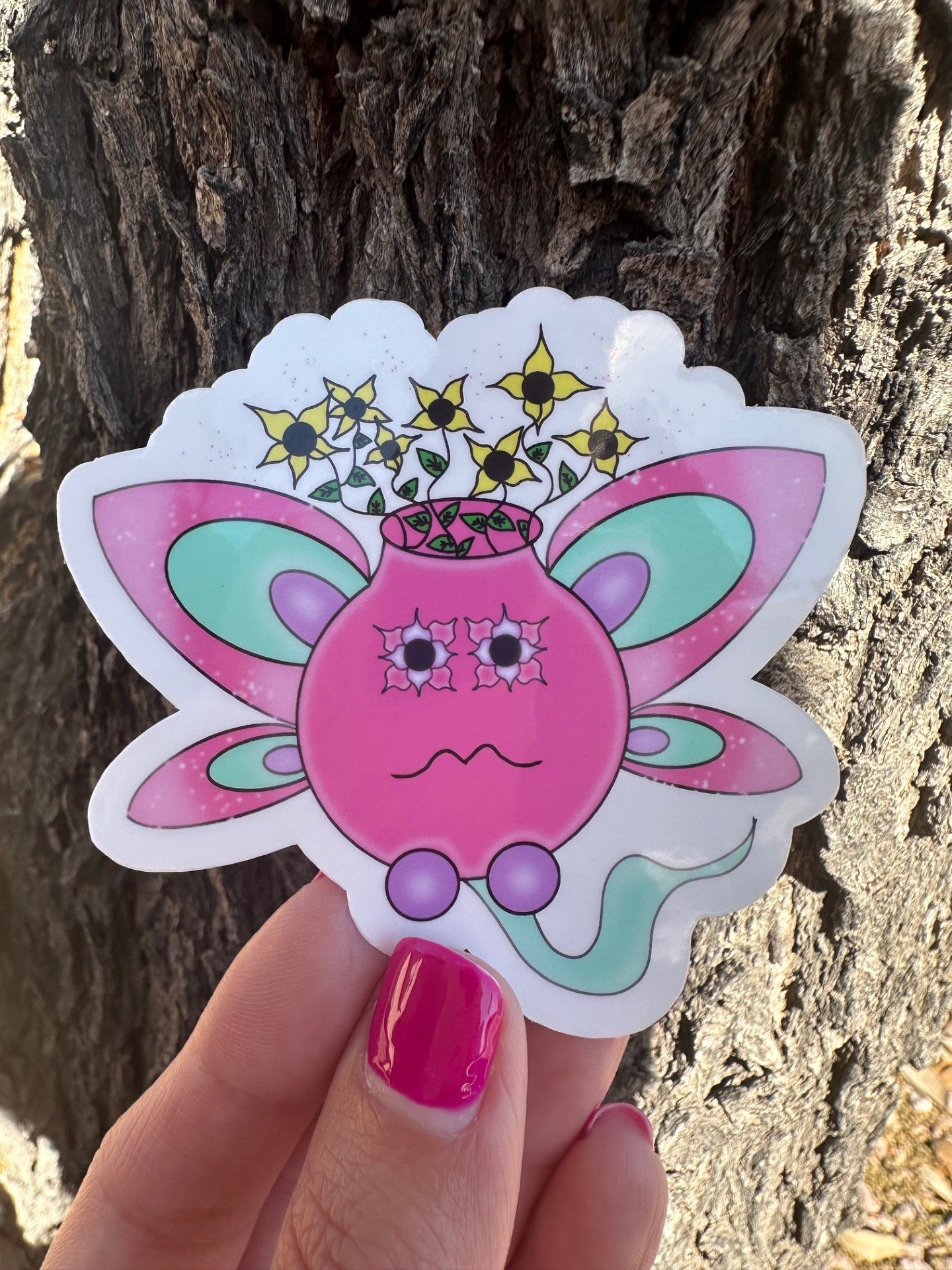A fictional character sticker that looks like a flower pot with wings coming out of it. The character is pink and turquoise with sparkles around it. The character has yellow flowers coming out of it. The sticker is held up against a tree branch. 