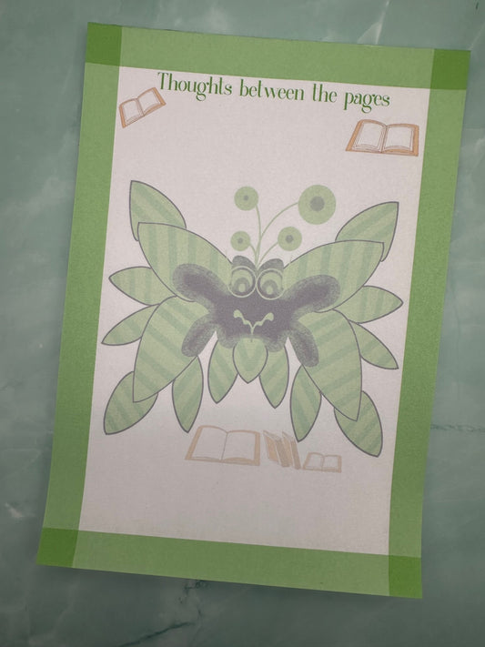 A notepad with fictional character that is green and black with black leaves coming from it with antennas. The character has green eyes with glasses. The notepad has green edges all around with a notepad heading of "Thoughts between the pages". The notepad is sitting on a light green background.