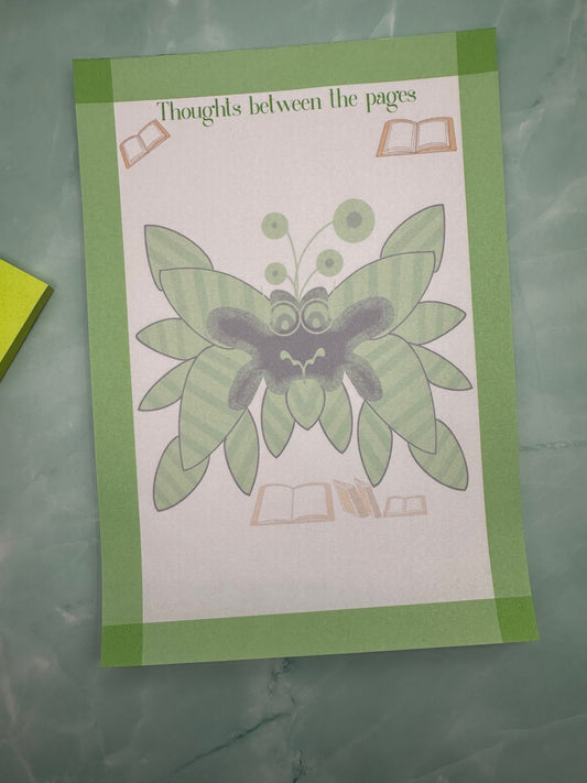 A notepad with fictional character that is green and black with black leaves coming from it with antennas. The character has green eyes with glasses. The notepad has green edges all around with a notepad heading of "Thoughts between the pages". The notepad is sitting on a light green background.
