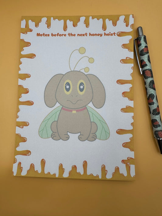 A notepad with a brown dog character that has yellow and black eyes with a red collar and green wings coming from it and also yellow antennas. All around the notepad is yellow honey with a notepad heading of "Notes before the next honey heist". The notepad is sitting on a yellow background.