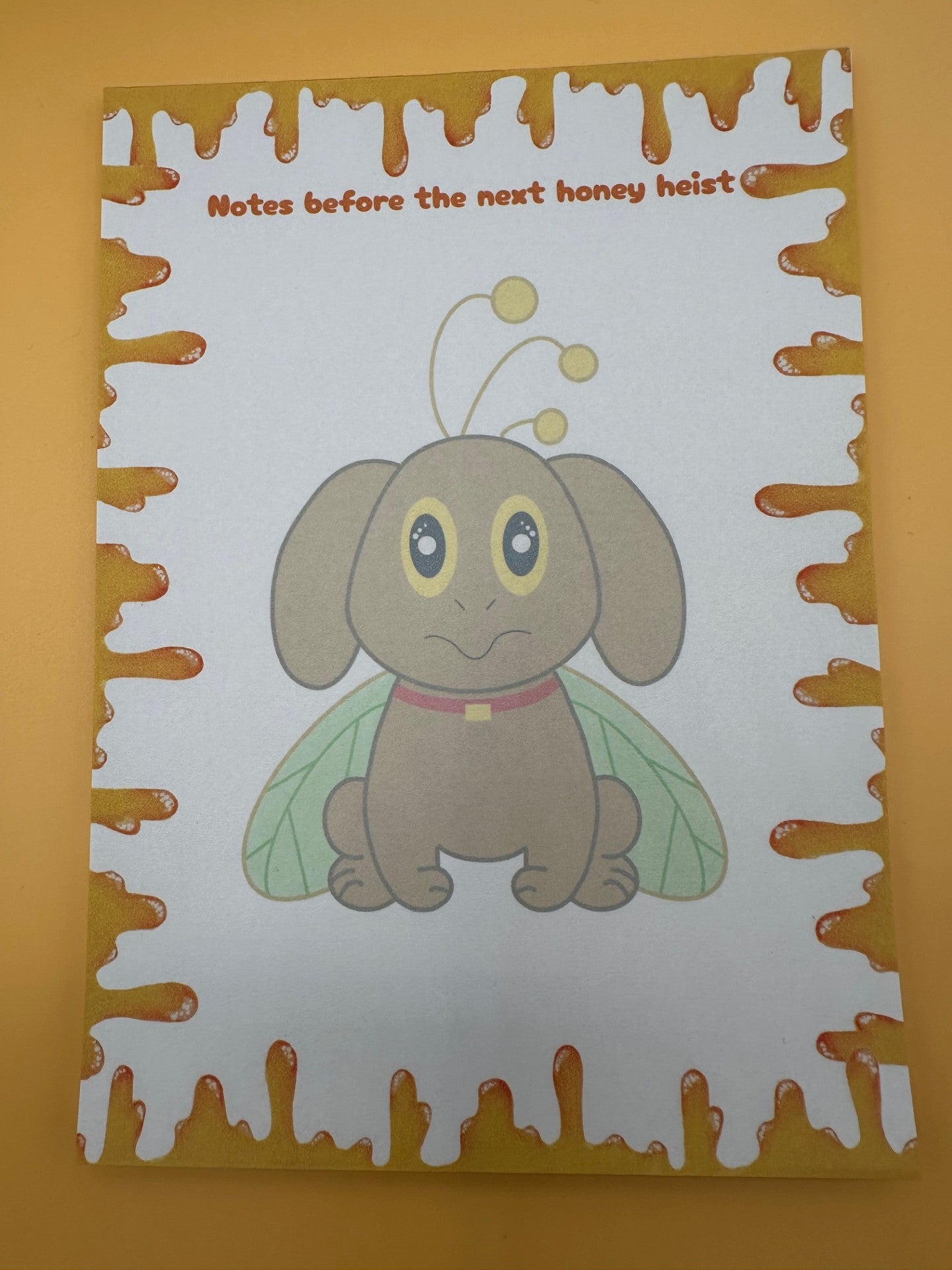 A notepad with a brown dog character that has yellow and black eyes with a red collar and green wings coming from it and also yellow antennas. All around the notepad is yellow honey with a notepad heading of "Notes before the next honey heist". The notepad is sitting on a yellow background.
