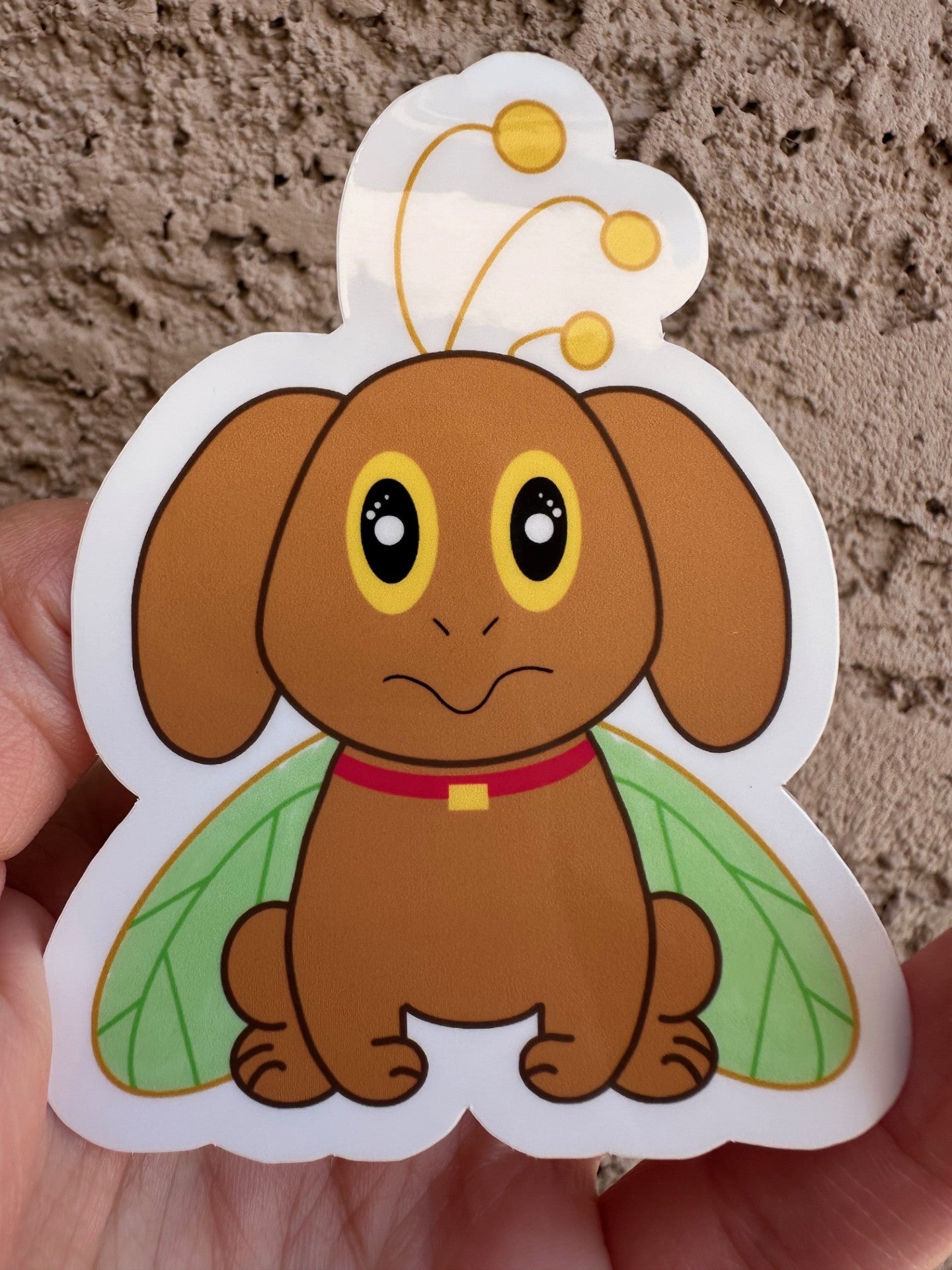 Cartoon brown dog that has big yellow and black eyes with green butterfly wings held against a textured beige background.