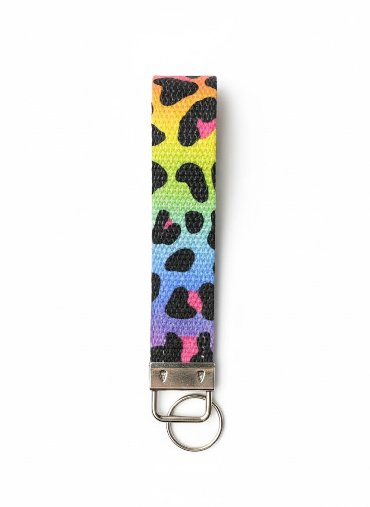 A fabric wristlet keychain with a rainbow gradient background in yellow, green, blue, and purple, featuring a black and pink leopard print pattern and a silver-toned metal key ring attachment.
