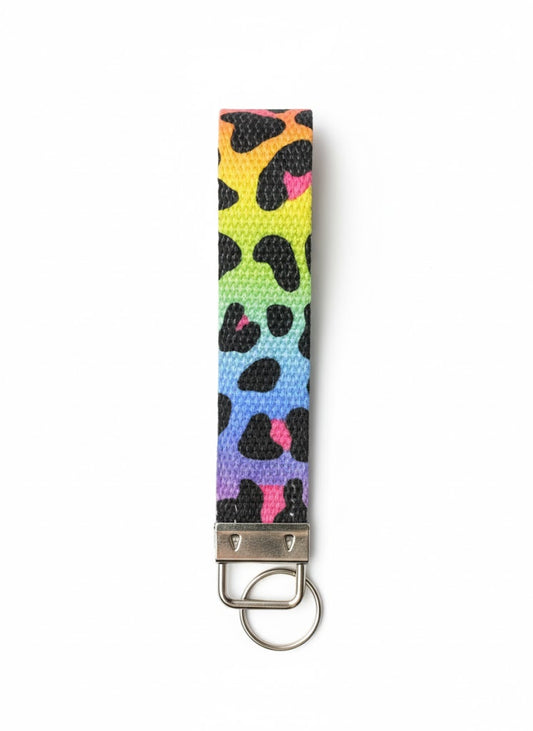 A fabric wristlet keychain with a rainbow gradient background in yellow, green, blue, and purple, featuring a black and pink leopard print pattern and a silver-toned metal key ring attachment.