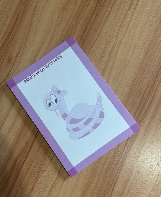 Notepad with a hand-drawn cartoon snake design that is purple, with a heading on the notepad that states "mind your business " on a wooden surface.
