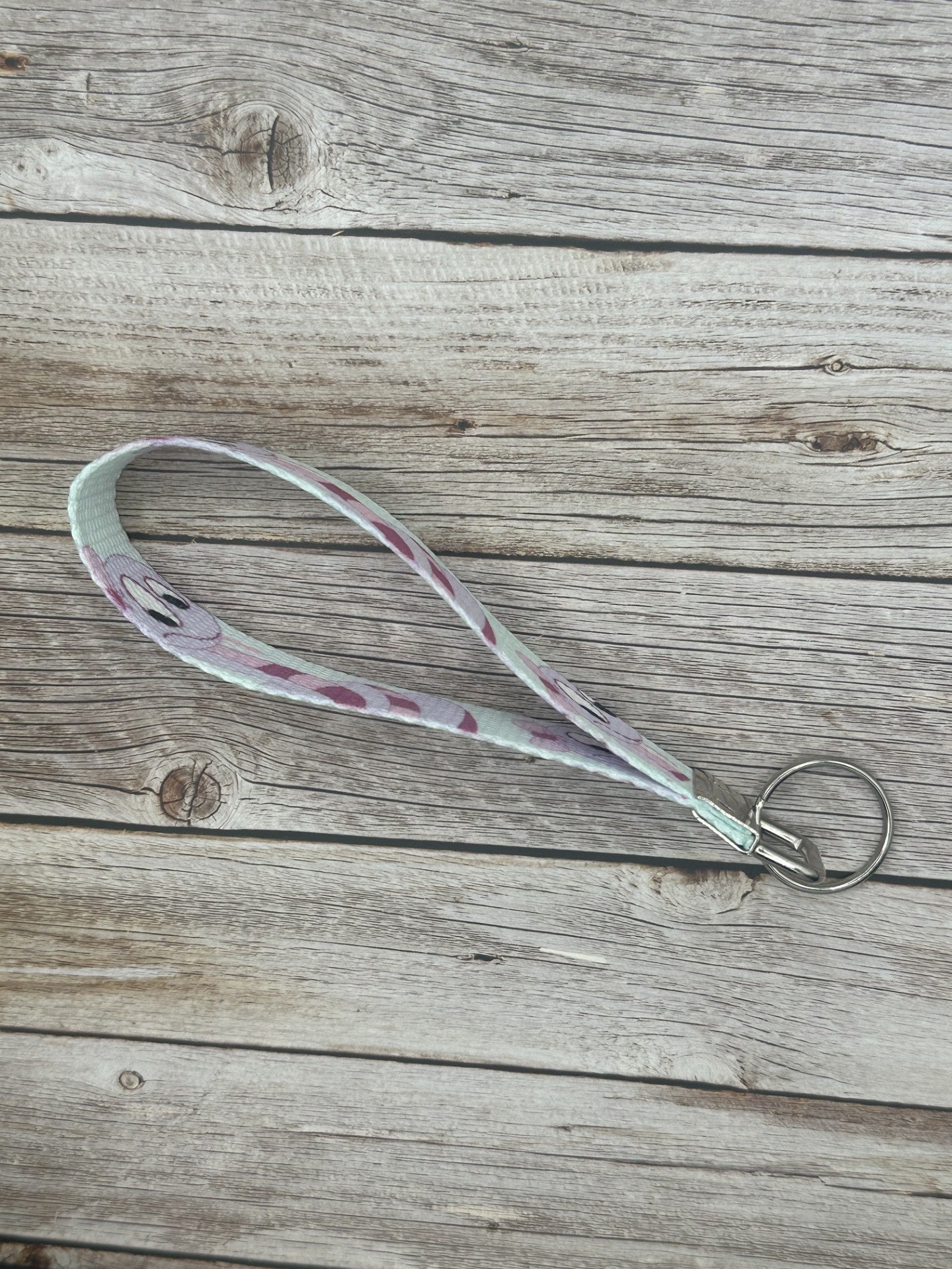 Keychain with a purple cartoon character that is a snake and has dark and light purple colors, with a bow on its head, the wristlet is on a wooden surface side view.