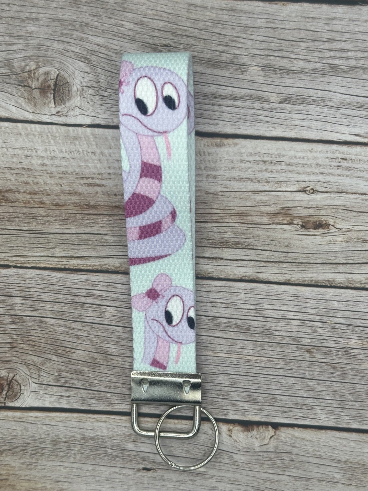 Keychain with a purple cartoon character that is a snake and has dark and light purple colors, with a bow on its head, the wristlet is on a wooden surface.