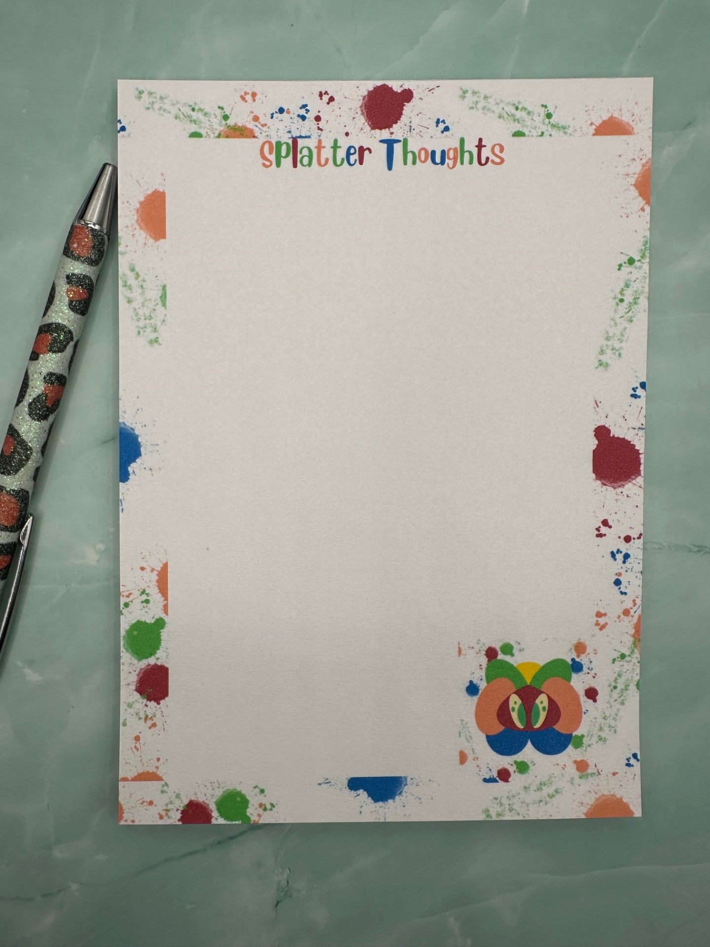 A notepad with a fictional character at the bottom of the notepad that is yellow, green, orange and blue color with green eyes. All around the notepad is different color splatters with a notepad heading of "Splatter thoughts". The notepad is sitting on a light green color background.  
