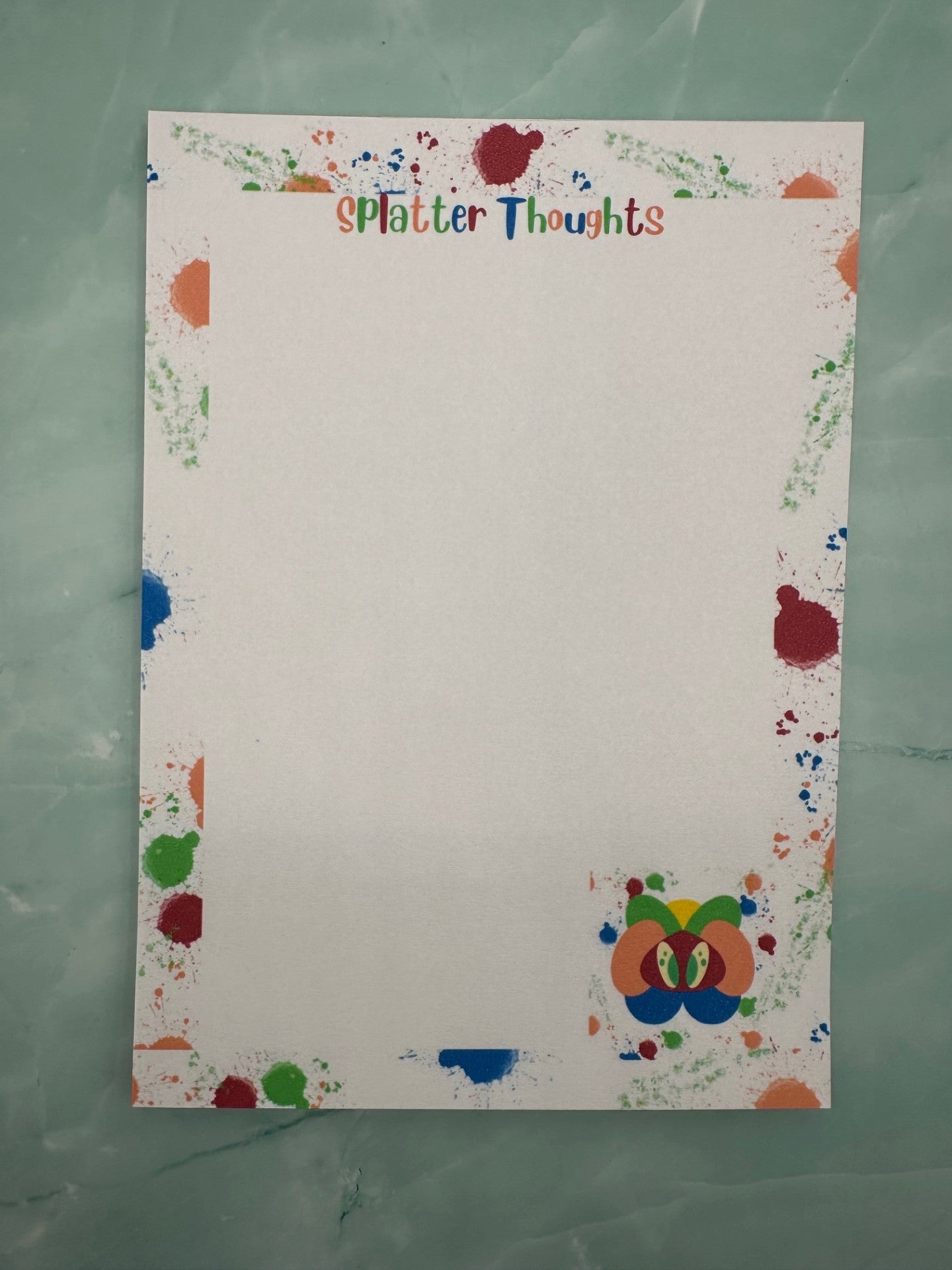 A notepad with a fictional character at the bottom of the notepad that is yellow, green, orange and blue color with green eyes. All around the notepad is different color splatters with a notepad heading of "Splatter thoughts". The notepad is sitting on a light green color background.  