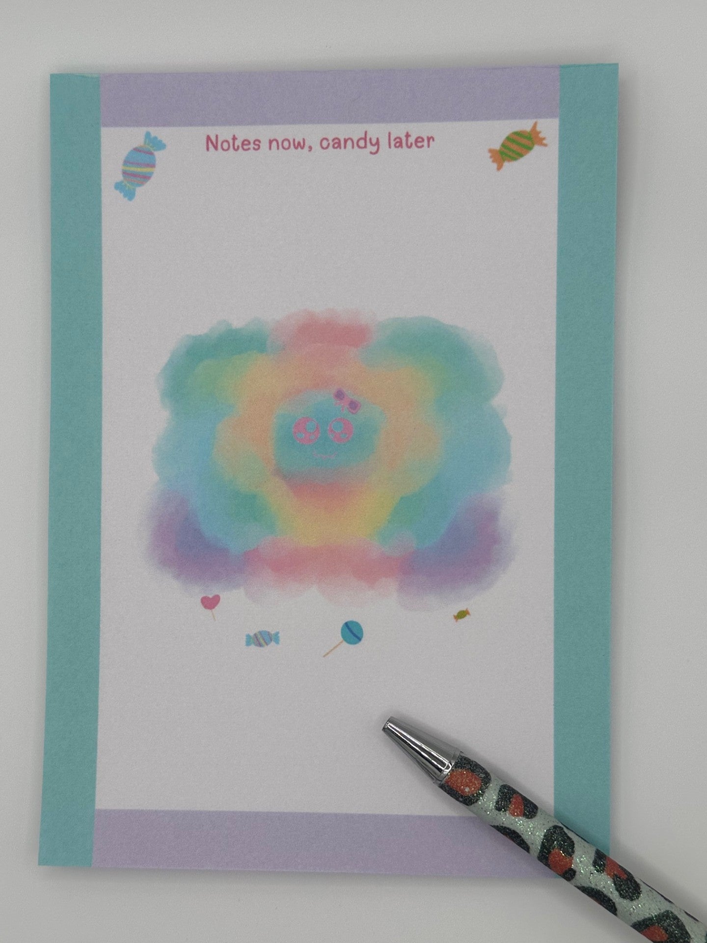 A notepad that has a pastel character that looks like a cloud. The character has a pink and purple bow with a pink eyes and smile. Candy is at the bottom of the cloudy pastel character and also on the top of the notepad. The notepad corner edges are light turquoise and light purple. The notepad heading "Notes now, candy later". The notepad is sitting on top of a white background.