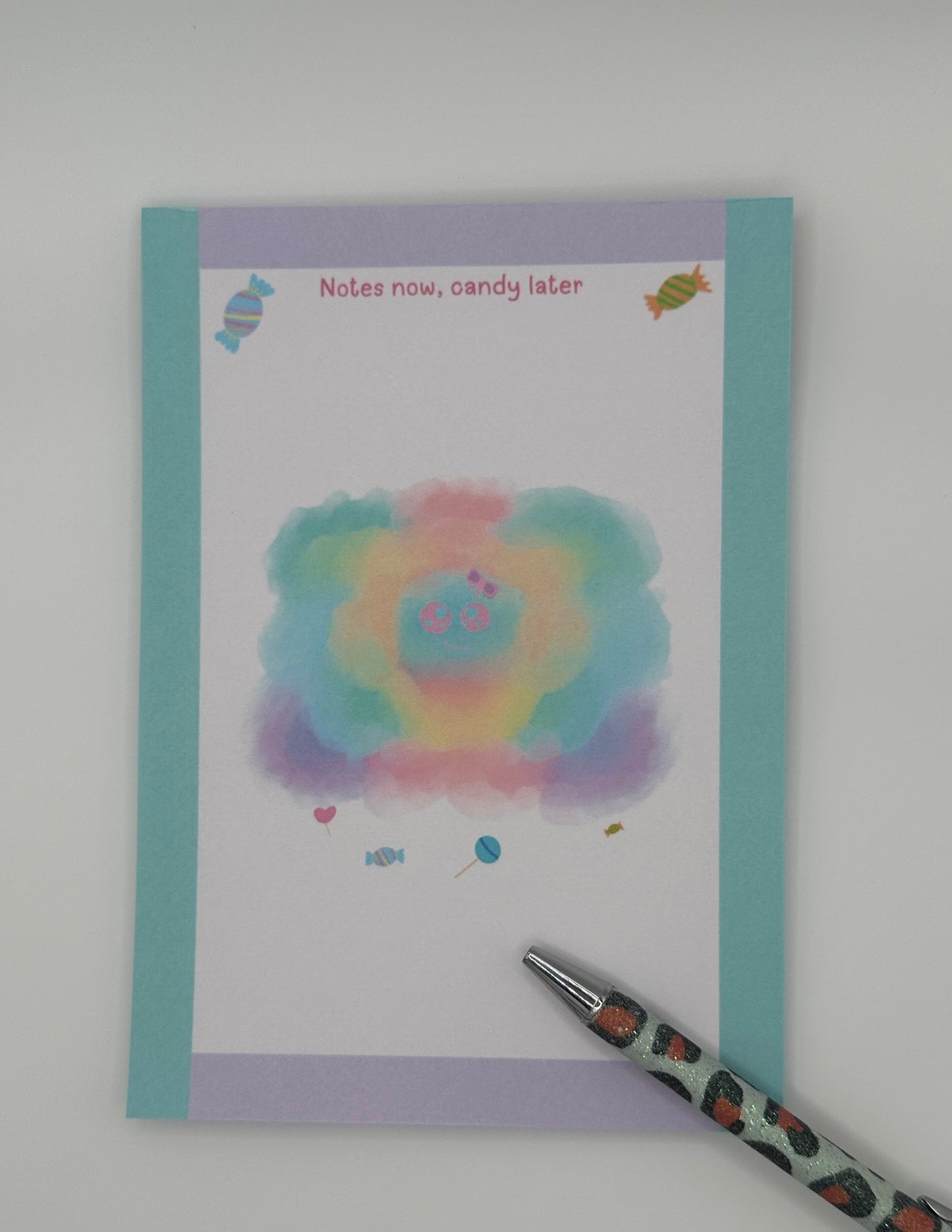 A notepad that has a pastel character that looks like a cloud. The character has a pink and purple bow with a pink eyes and smile. Candy is at the bottom of the cloudy pastel character and also on the top of the notepad. The notepad corner edges are light turquoise and light purple. The notepad heading "Notes now, candy later". The notepad is sitting on top of a white background.