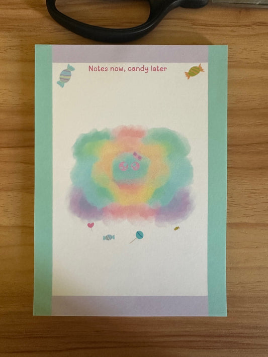 Colorful character design "sprinkles" that has different pastel colors with candy beneath the character. The notepad is sitting on wooden surface with a heading on the notepad "notes now, candy later".
