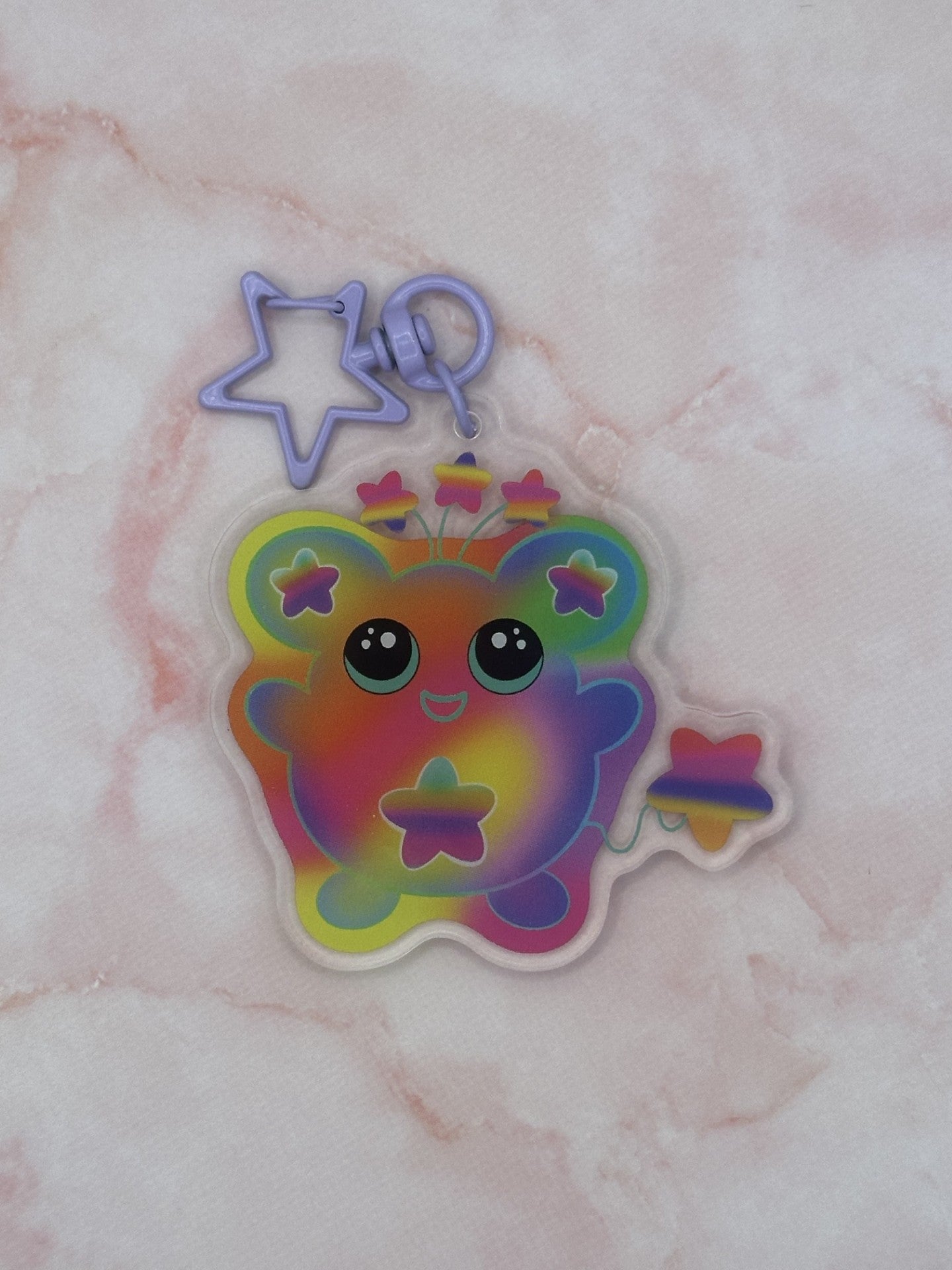 Colorful character that is different rainbow colors that has a star on its stomach and stars on the ears. The character has a colorful tail as well. The eyes are black with turquoise in them. The character also has three stars coming out on top of its head. The keychain has a light purple keyring. 