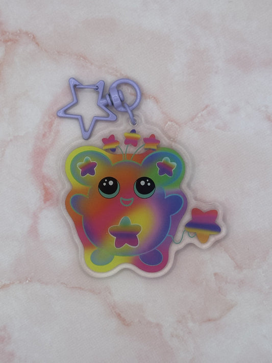 Colorful character that is different rainbow colors that has a star on its stomach and stars on the ears. The character has a colorful tail as well. The eyes are black with turquoise in them. The character also has three stars coming out on top of its head. The keychain has a light purple keyring. 