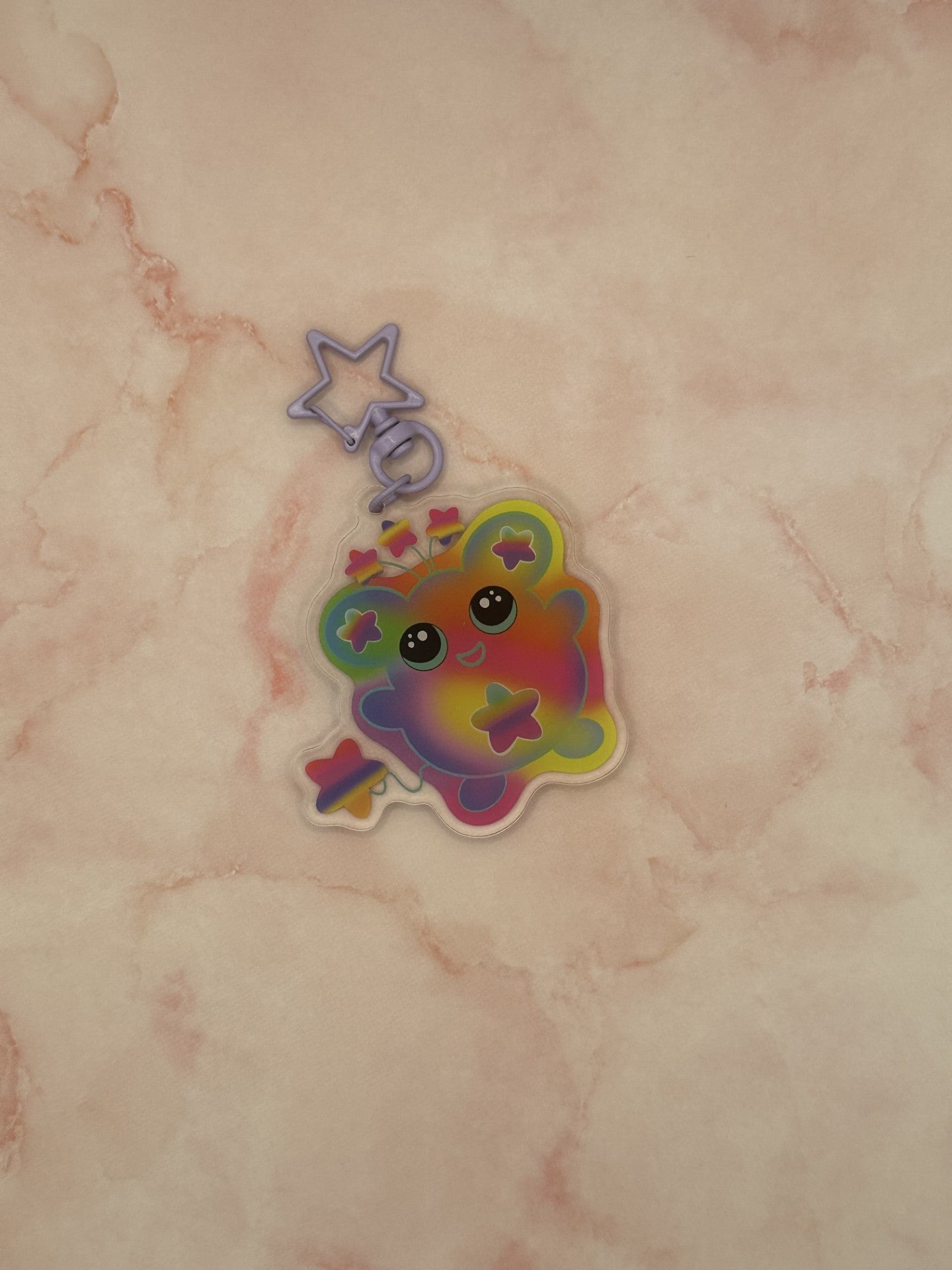 Colorful character that is different rainbow colors that has a star on its stomach and stars on the ears. The character has a colorful tail as well. The eyes are black with turquoise in them. The character also has three stars coming out on top of its head. The keychain has a light purple keyring. 