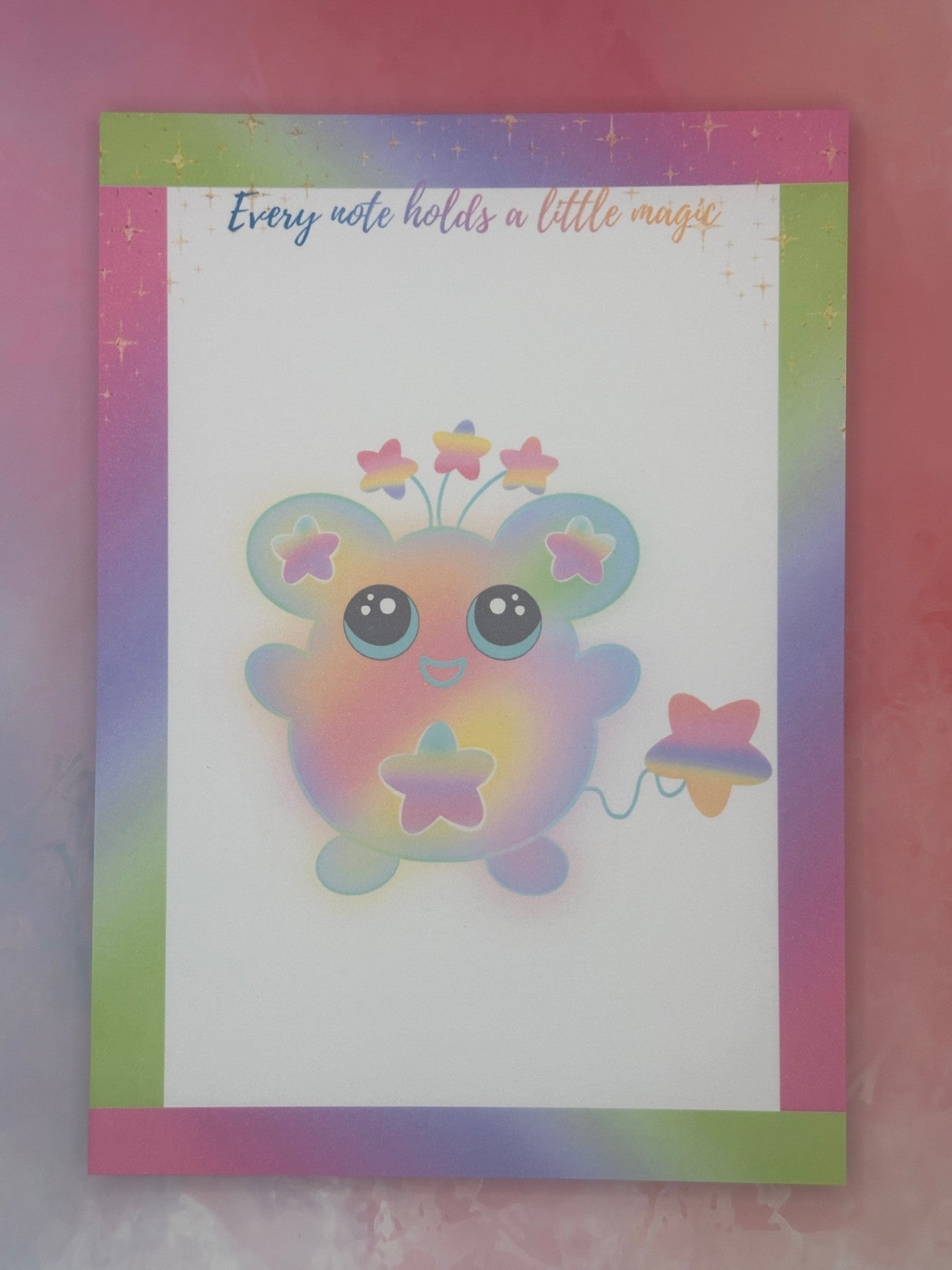 Notepad that has a colorful cartoon character that has stars on the ears and a star on the stomach. The character has different colors like pink, purple, green, and yellow. The character has stars coming from outside of its head and has a star as a tail. The notepad has neon bright colors on all four corners and has sparkling stars on top of the notepad. The notepad has the heading "Every note holds a little magic". 