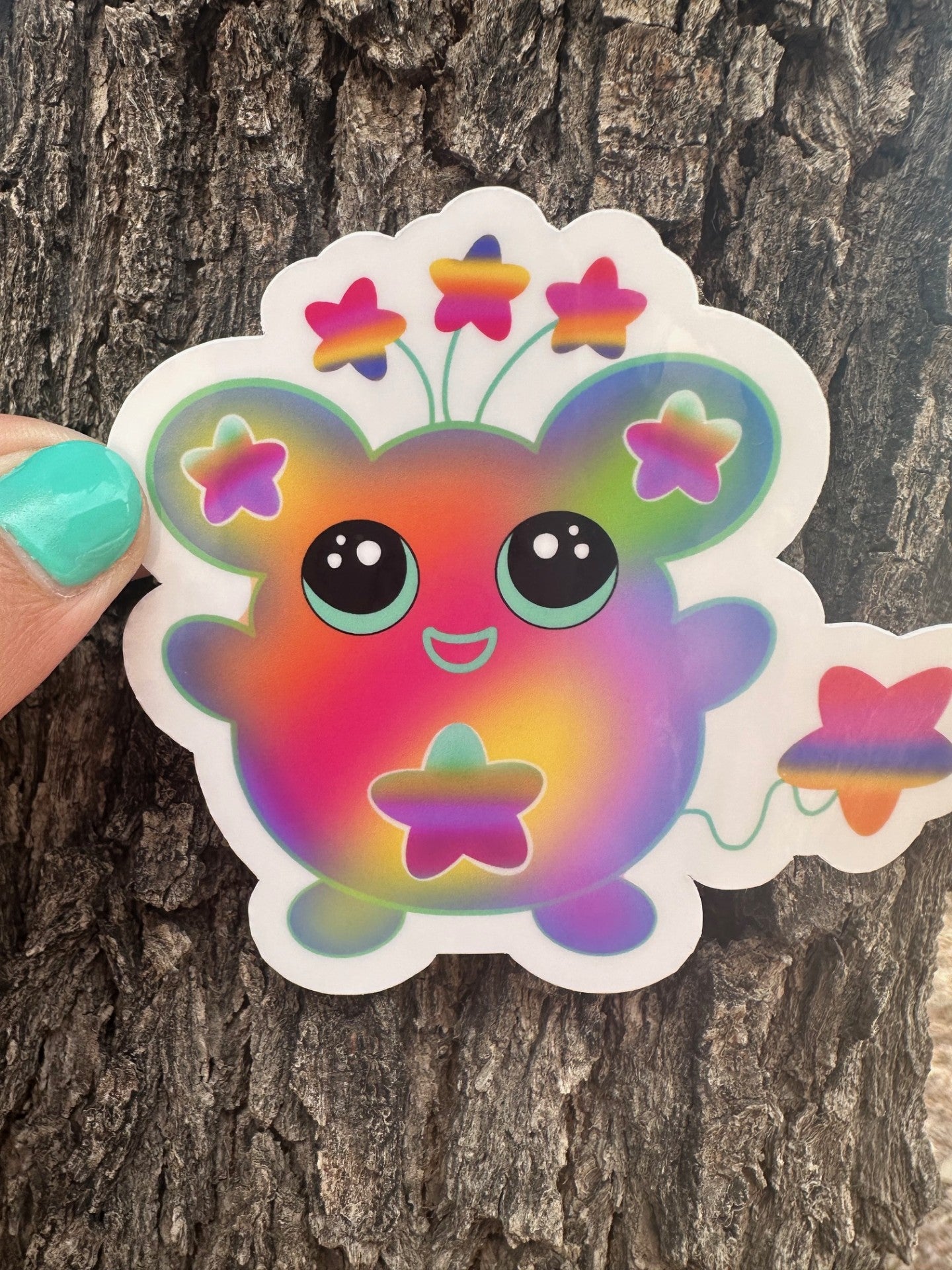Colorful cartoon character sticker that has stars on the ears and a star on the stomach. The character has different colors like pink, purple, green, and yellow. The character has stars coming from outside of its head and has a star as a tail. The character sticker design is held up against a tree trunk. 
