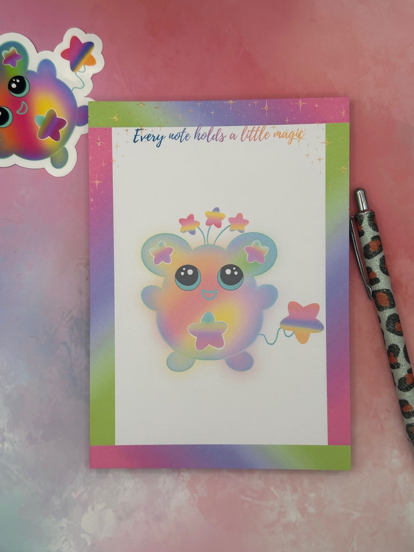 Notepad that has a colorful cartoon character that has stars on the ears and a star on the stomach. The character has different colors like pink, purple, green, and yellow. The character has stars coming from outside of its head and has a star as a tail. The notepad has neon bright colors on all four corners and has sparkling stars on top of the notepad. The notepad has the heading "Every note holds a little magic". The notepad is sitting on a colorful background with a pen and a sticker.