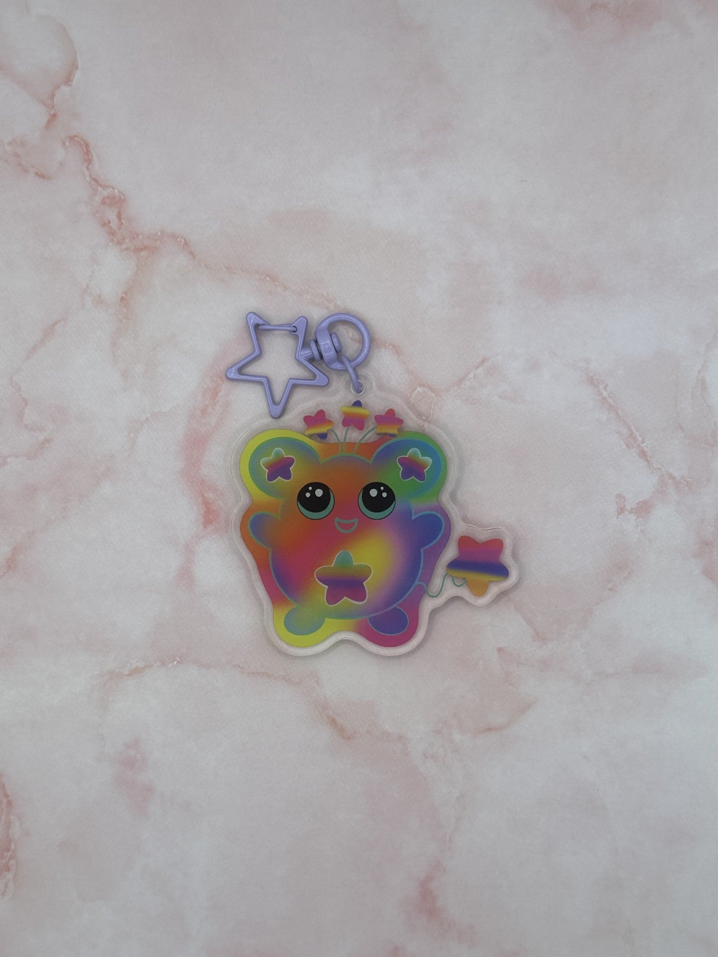 Colorful character that is different rainbow colors that has a star on its stomach and stars on the ears. The character has a colorful tail as well. The eyes are black with turquoise in them. The character also has three stars coming out on top of its head. The keychain has a light purple keyring. 