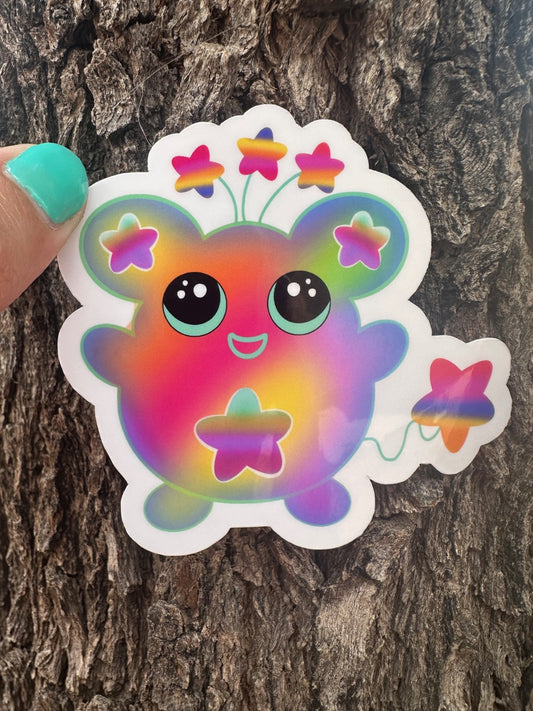 Colorful cartoon character sticker that has stars on the ears and a star on the stomach. The character has different colors like pink, purple, green, and yellow. The character has stars coming from outside of its head and has a star as a tail. The character sticker design is held up against a tree trunk. 