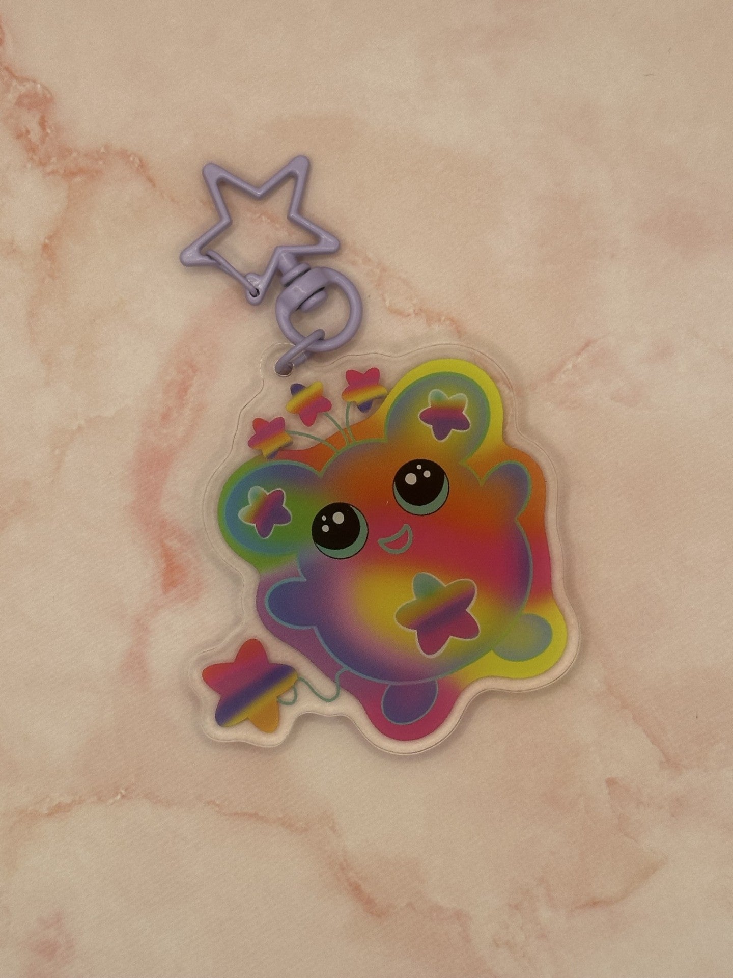 Colorful character that is different rainbow colors that has a star on its stomach and stars on the ears. The character has a colorful tail as well. The eyes are black with turquoise in them. The character also has three stars coming out on top of its head. The keychain has a light purple keyring. 