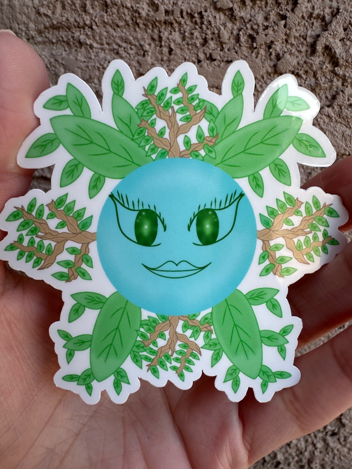 Glossy sticker of fictional character that is blue circle shape with green face coming out of it, has green leaves and branches. 