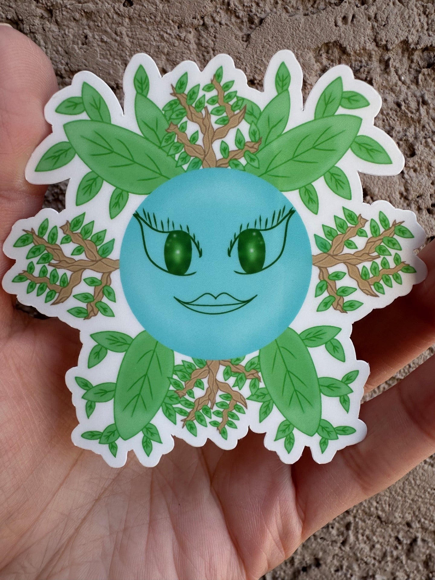 Glossy sticker of a fictional character shaped like a blue circle with green eyes and a green mouth, coming out of which are green leaves and branches. 