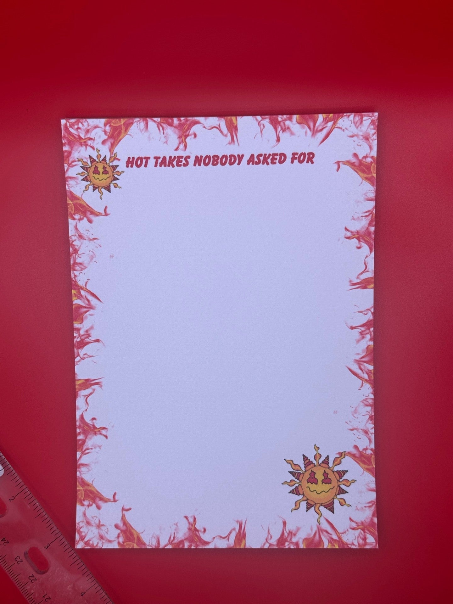 A notepad with character on top and bottom of notepad that looks like a sun with yellow and red colors and a smirk face. The notepad edges all around the corners have orange and red flames with the notepad heading of "Hot takes nobody asked for". The notepad is sitting on a red background next to a ruler. 
