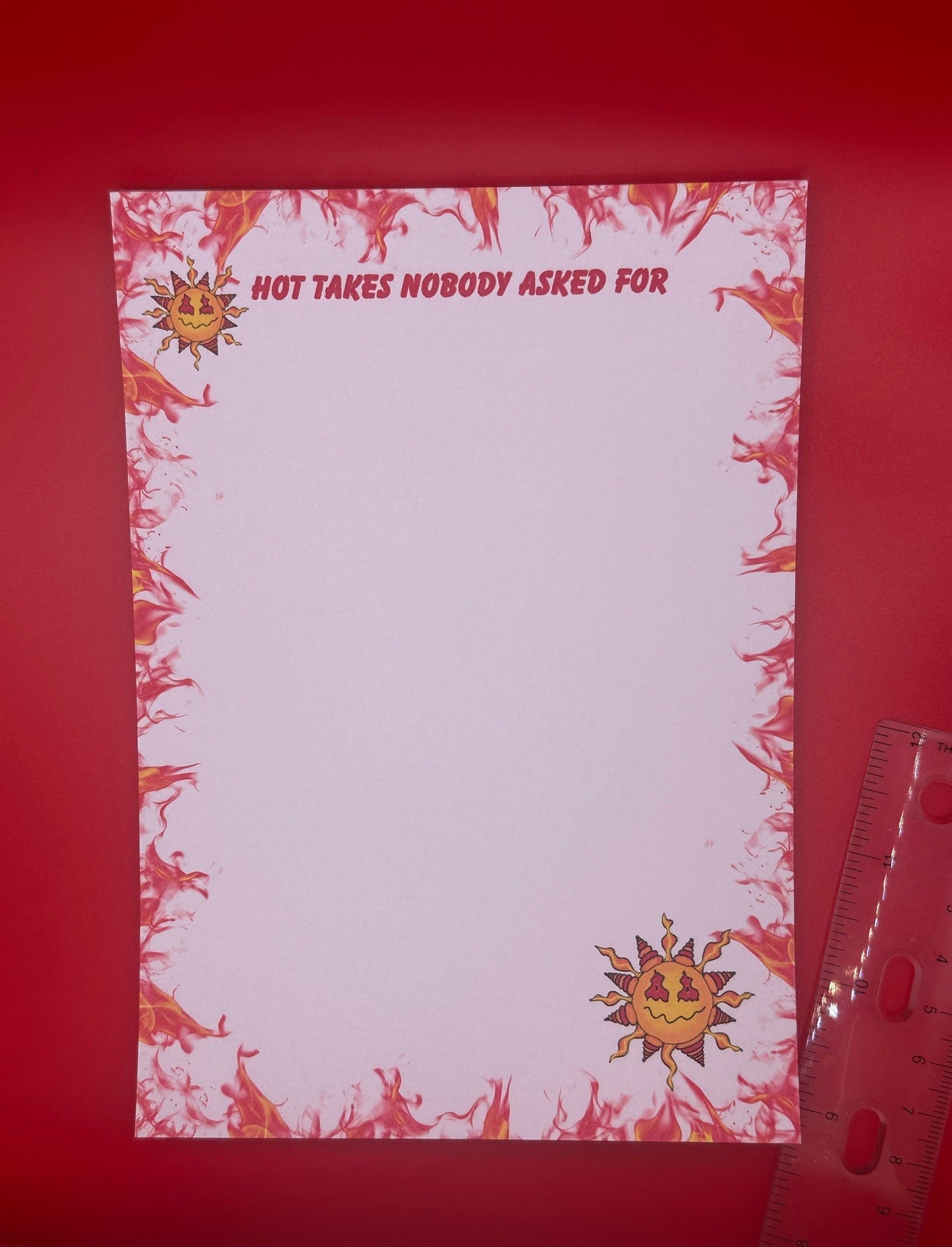 A notepad with character on top and bottom of notepad that looks like a sun with yellow and red colors and a smirk face. The notepad edges all around the corners have orange and red flames with the notepad heading of "Hot takes nobody asked for". The notepad is sitting on a red background next to a ruler. 