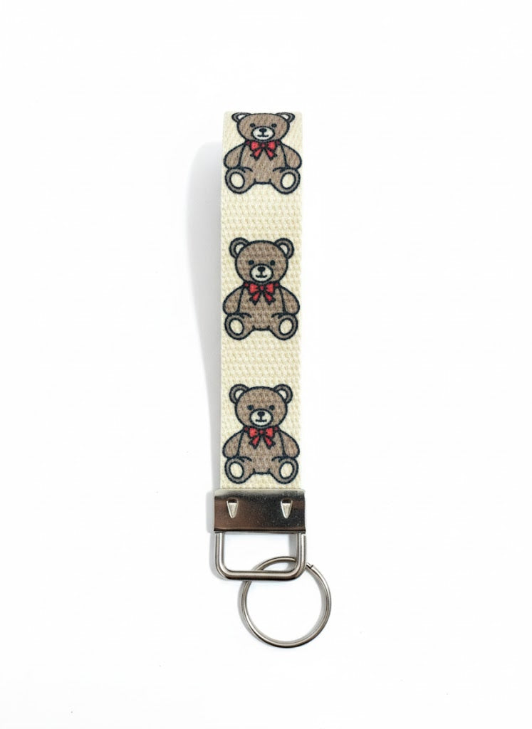 A fabric wristlet keychain with a cream background, patterned with three embroidered brown teddy bears wearing red bows.