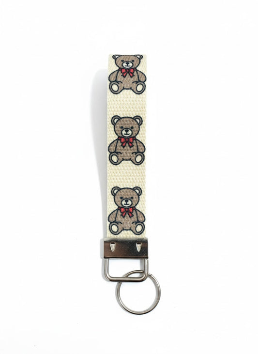 A fabric wristlet keychain with a cream background, patterned with three embroidered brown teddy bears wearing red bows.