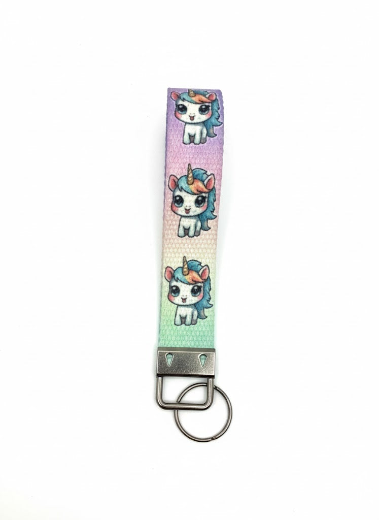 A fabric wristlet keychain with a purple, blue, and green gradient background, patterned with cartoon unicorns and featuring a silver-toned metal key ring attachment.