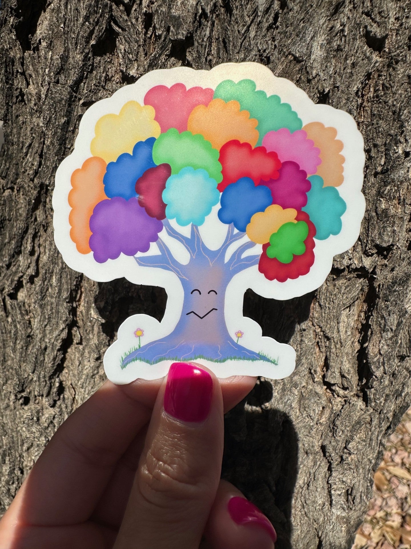 Rainbow-colored tree sticker, with different vibrant colors, with black eyes and a mouth, and is held in front of the tree bark. 