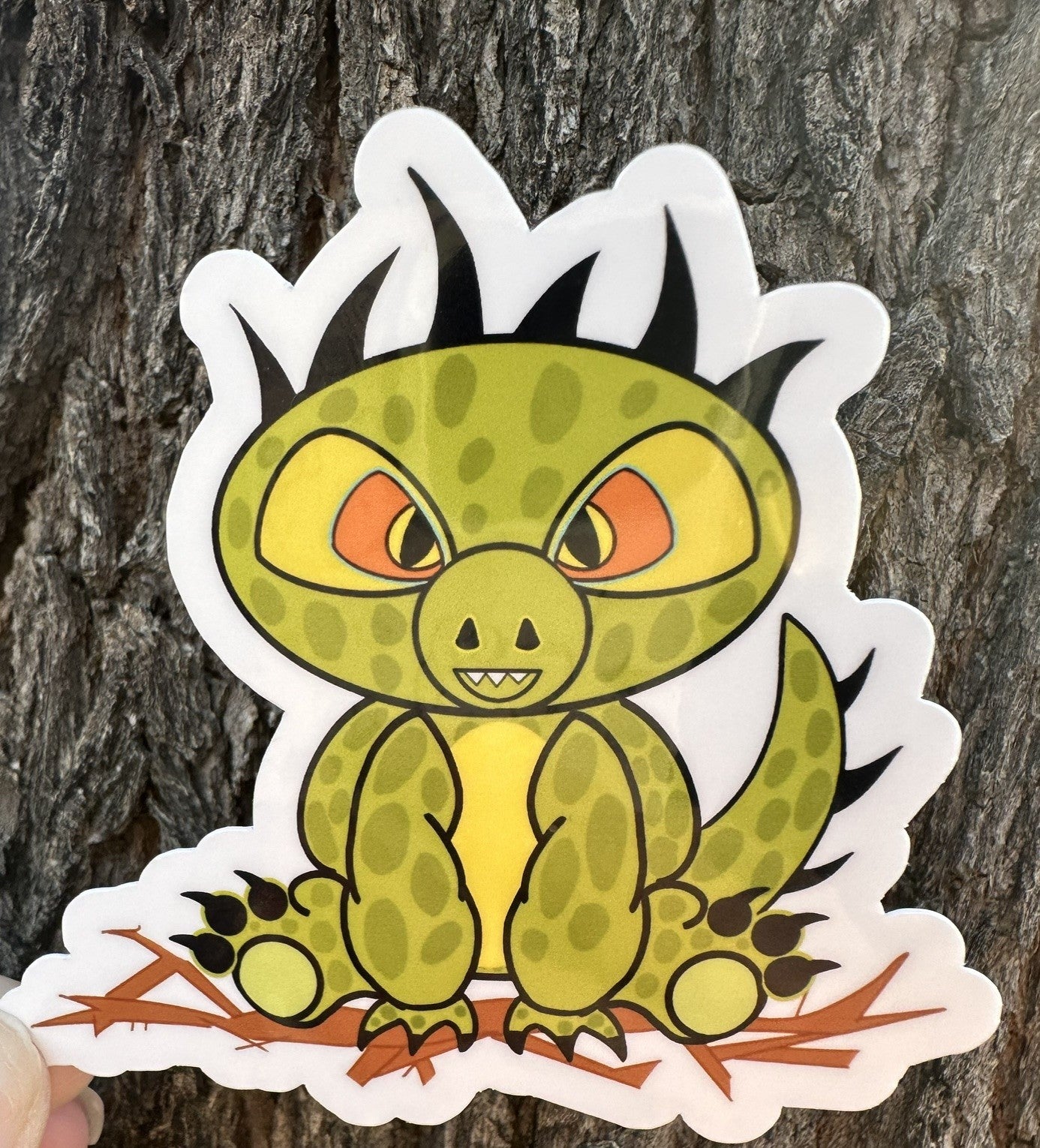 Cartoon half dinosaur half lizard sticker. The sticker character is green and light lime in the middle of its stomach. The character also has dark green dots with black hair and has black claws coming from the hands and feet. The character has orange and black eyes. The character is sitting on top of brown branches. The sticker background is a tree.