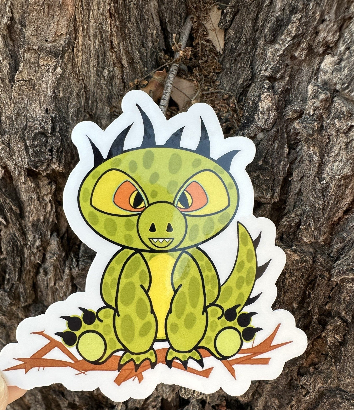 Cartoon half dinosaur half lizard sticker. The sticker character is green and light lime in the middle of its stomach. The character also has dark green dots with black hair and has black claws coming from the hands and feet. The character has orange and black eyes. The character is sitting on top of brown branches. The sticker background is a tree.